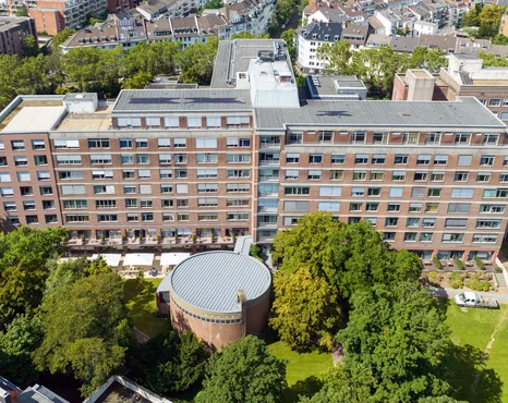 Luftaufnahme eines großen, mehrstöckigen Backsteingebäudes mit einem runden Bauwerk davor, umgeben von Bäumen und grünen Rasenflächen sowie nahegelegenen städtischen Gebäuden.
