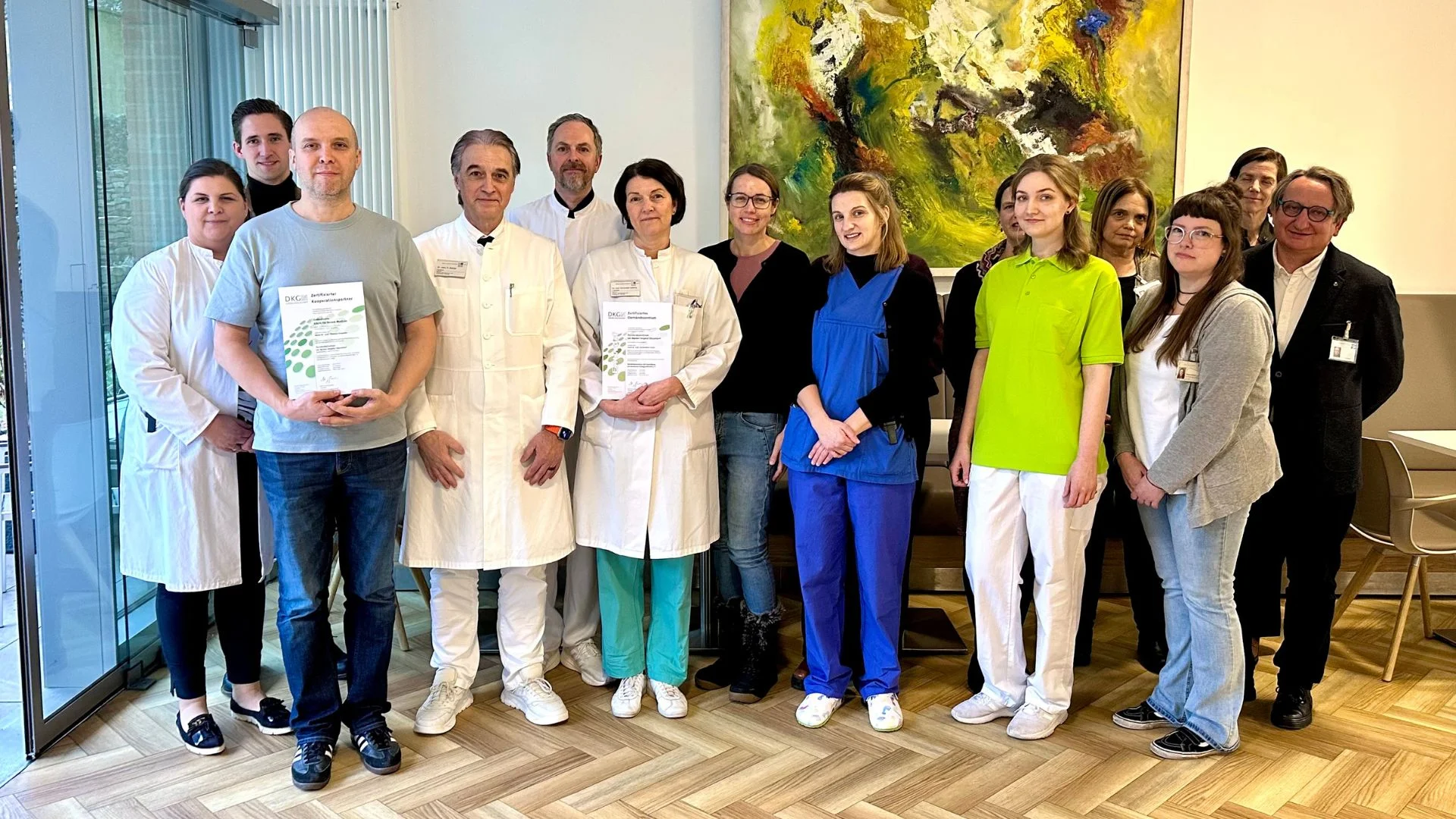 Dreizehn Personen, darunter sowohl solche in medizinischer Uniform als auch in Freizeitkleidung, stehen gemeinsam in einem Raum auf einem Holzboden für ein Gruppenfoto. Drei von ihnen halten Urkunden in den Händen. Hinter ihnen hängt ein farbenfrohes abstraktes Gemälde an der Wand.